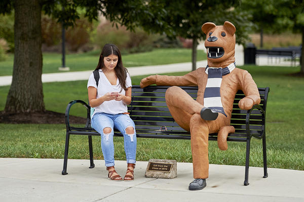 Spending time with the Nittany Lion A girl checks her phone while seated on a bench next to the Nittany Lion mascot.