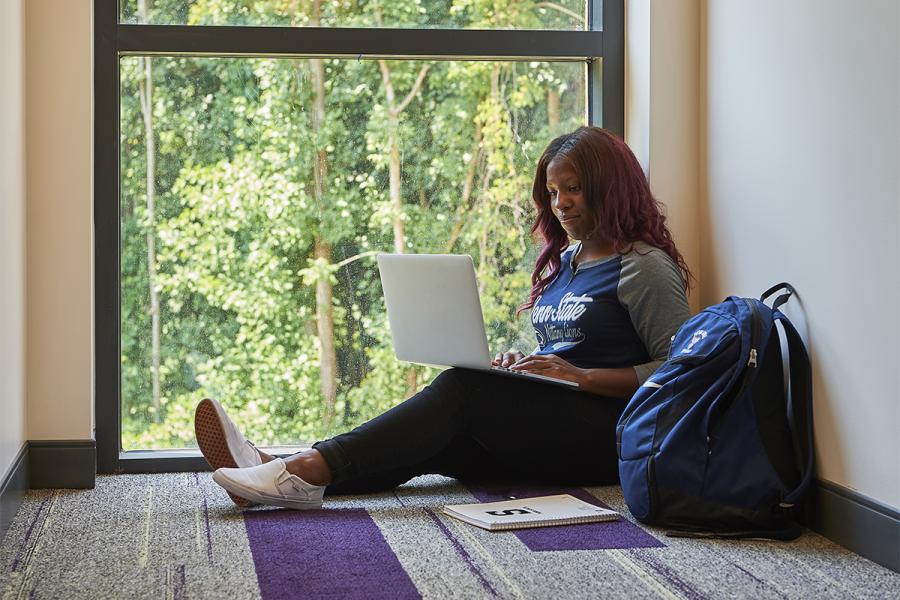 Student on laptop next to a window in the hallway