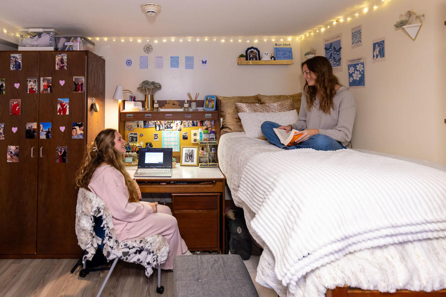 Inside a dorm bedroom, two girls sit across from one another chatting. One sits on top of a bed with white sheets and the other sits at the desk. 