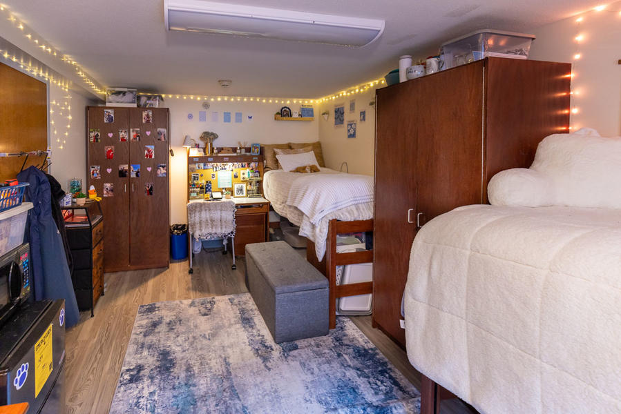 Two dorm beds placed parallel against the wall of a dorm bedroom on the right side. On the left said there are other amenities such as a wardrobe and desk. The room is lined with string lights. 