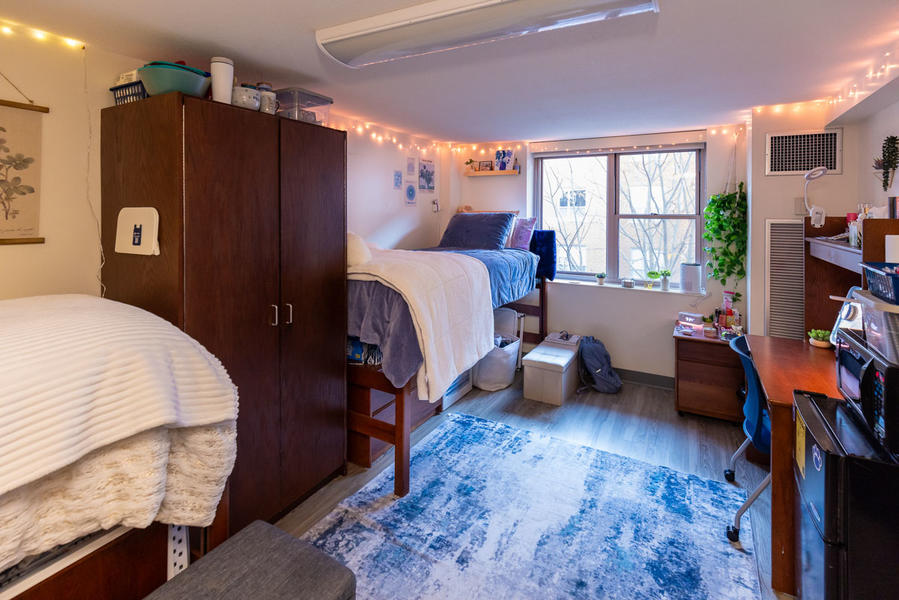 Two dorm beds placed parallel against the wall of a dorm bedroom. On the far wall, there is a window letting in light. The room is lined with string lights. 