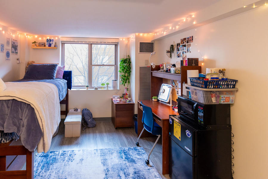 Inside a dorm bedroom, there is a bed placed on the left side. Straight ahead on the far wall is a window. On the right side of the room there is a mircowave placed on a mini fridge alongside a desk. 