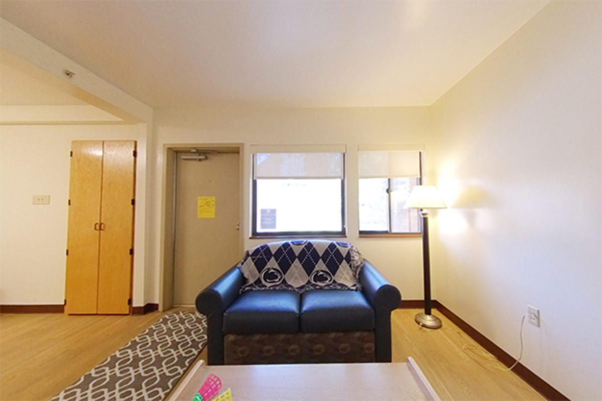 A bright living room with a blue sofa draped with a Nittany Lion blanket, wooden floor, and lamp.
